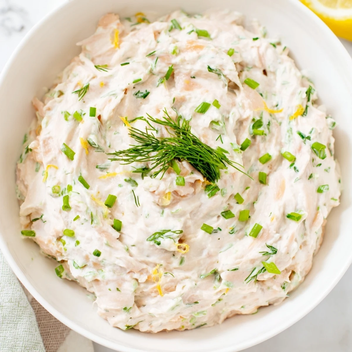 Smoked salmon dip in a white serving bowl, garnished with chives and lemon zest beside crunchy crackers.  