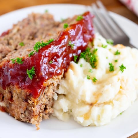 Golden-brown classic meatloaf topped with tangy glaze beside smooth mashed potatoes.  