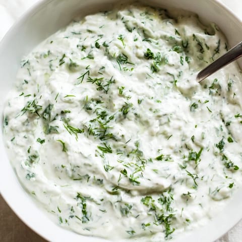 A chilled bowl of Greek Yogurt Herb Dip features vibrant green herbs and lemon zest, served with crunchy cucumber slices.  