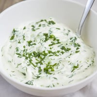 Creamy Greek Yogurt Herb Dip is spooned into a white bowl, surrounded by fresh vegetables and pita chips for dipping.  